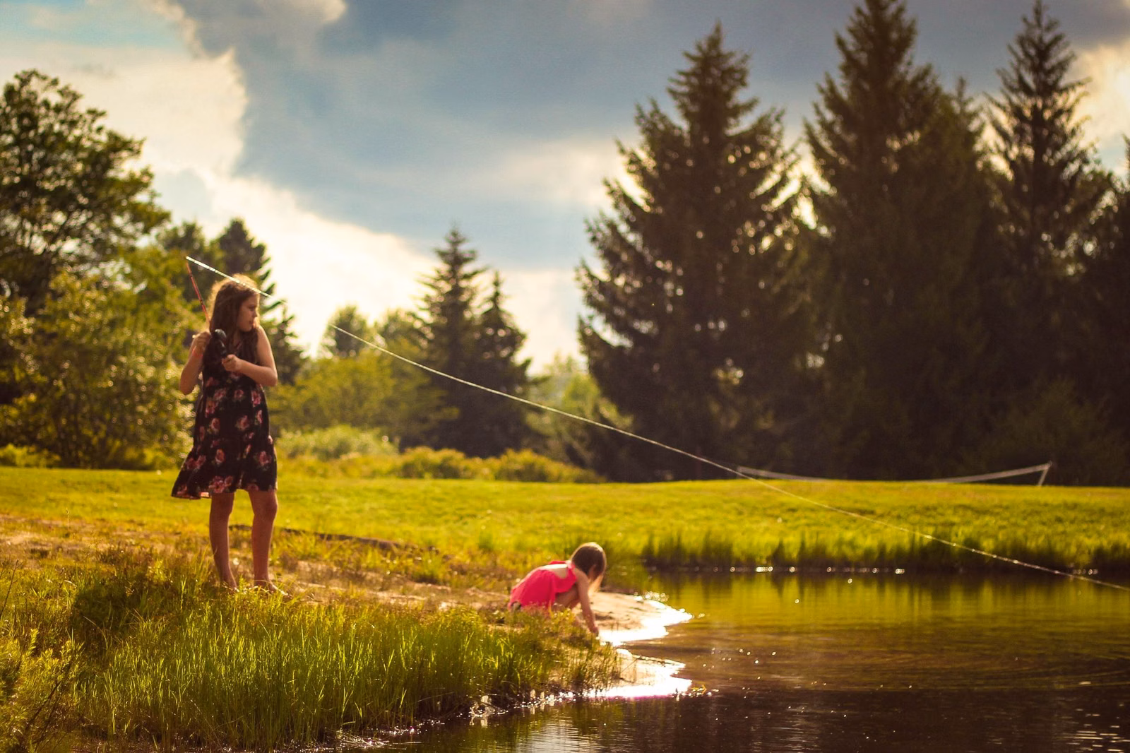Two children fishing from the grassy edge of a pond with tall evergreens behind